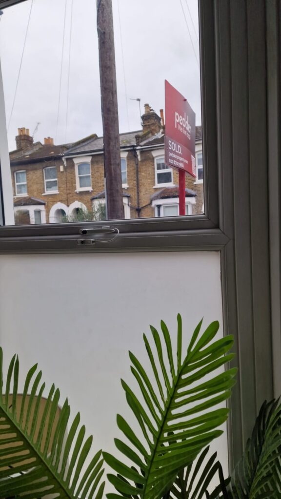 Living Room & Bedroom Renovation | London & Home Counties 17 A sold sign displayed outside a residential property viewed through a window with a plant in the foreground. - London