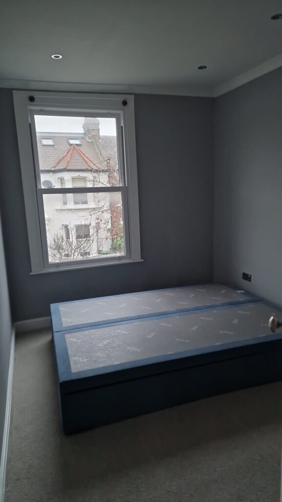 Living Room & Bedroom Renovation | London & Home Counties 19 A blue base bed with a mattress covered in plastic in a room with grey walls and a window. - London