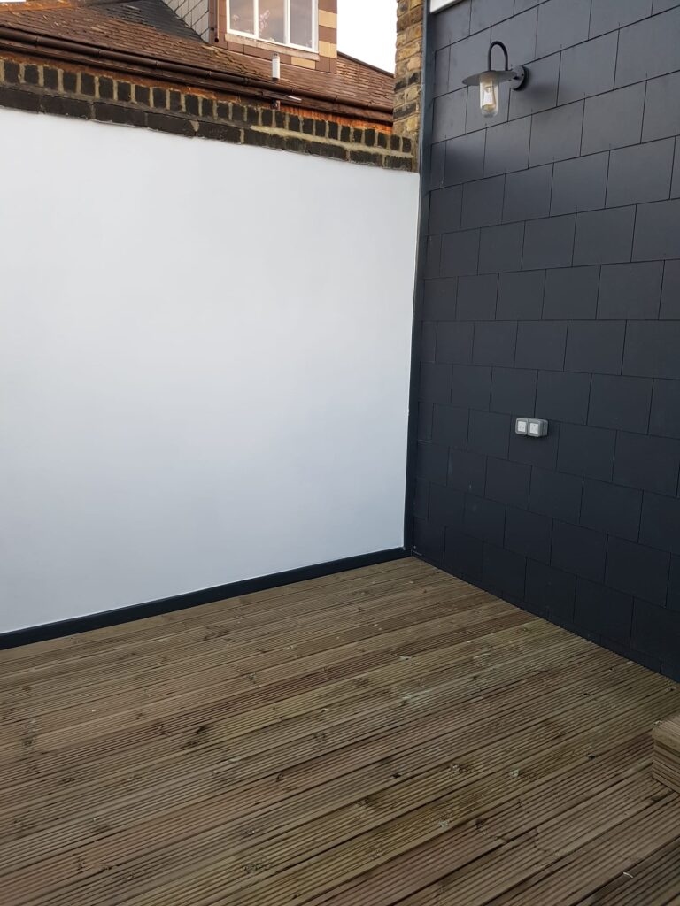 Garden Buildings & Outdoor Spaces | London & Home Counties 2 Wooden decking area adjacent to a grey wall with a light fixture and a blank white wall. - London