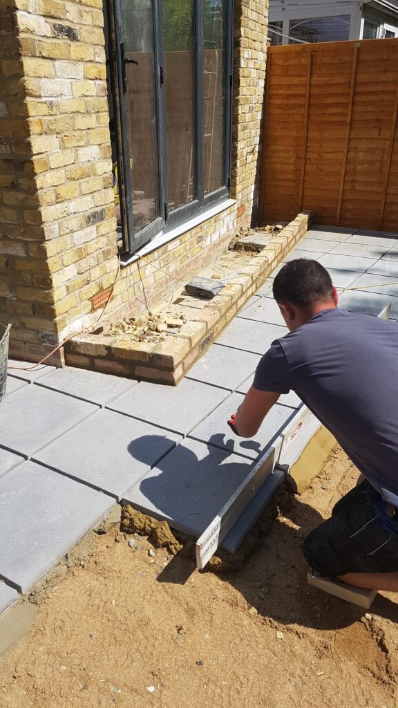 Garden Buildings & Outdoor Spaces | London & Home Counties 4 Individual installing grey paving stones in a garden area with a brick wall in the background. - London