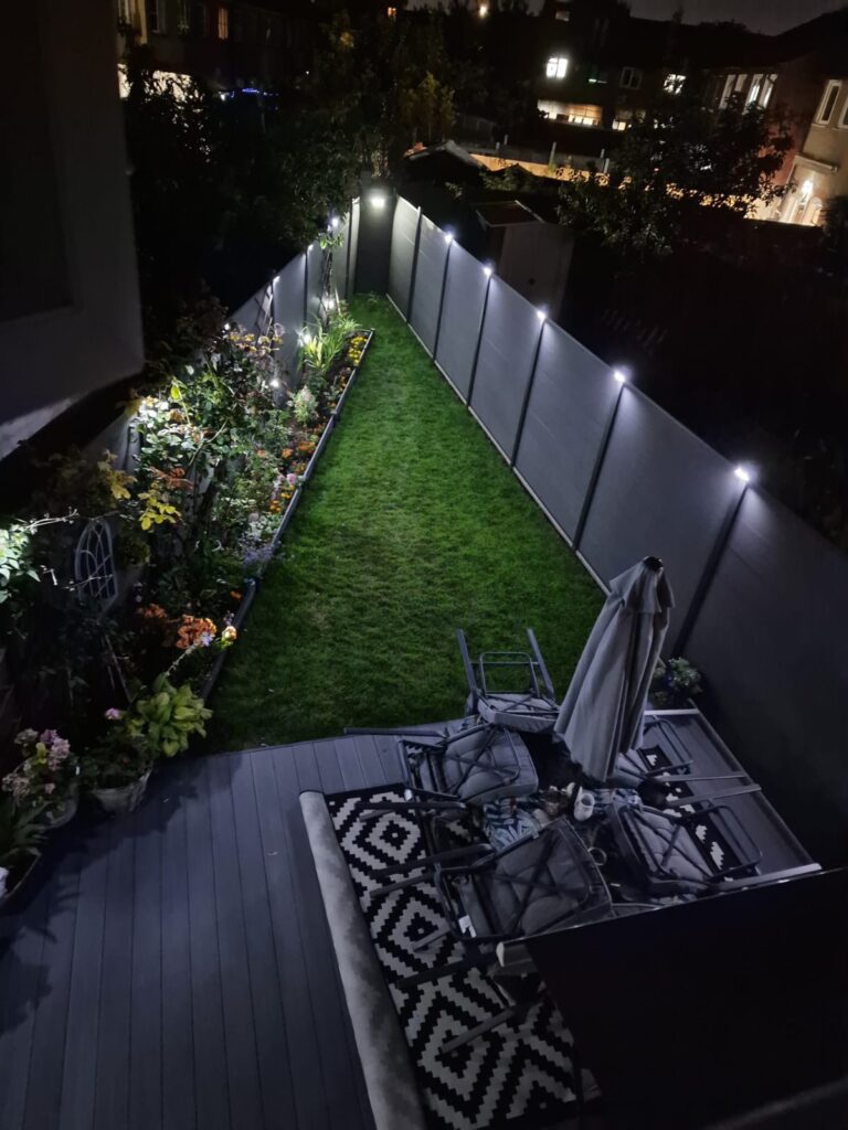 Garden Buildings & Outdoor Spaces | London & Home Counties 35 A garden at night featuring a well-lit pathway, seating area, and flower beds along the fence.